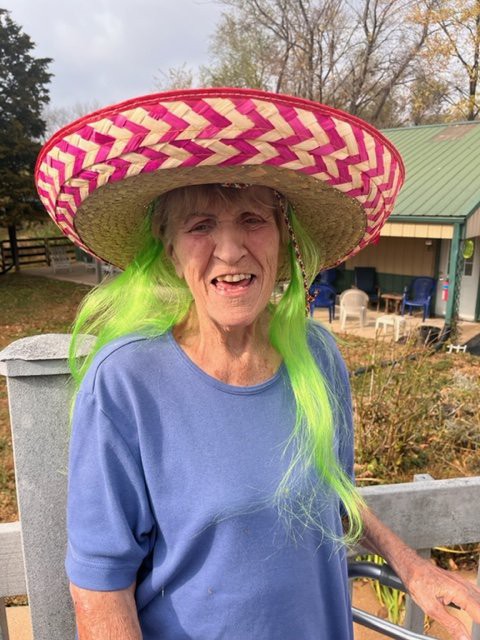 A woman wearing a colorful sombrero and green hair stands outdoors, smiling.