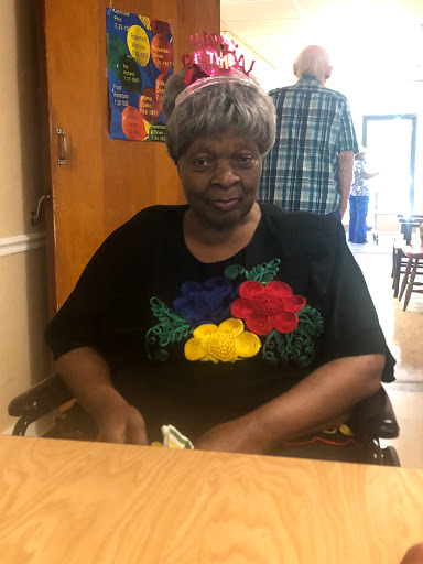 An elderly woman in a colorful shirt smiles while seated in a wheelchair, enjoying her time indoors.