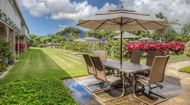 A scenic patio setup with outdoor furniture and an umbrella, overlooking a lush garden and lawn.