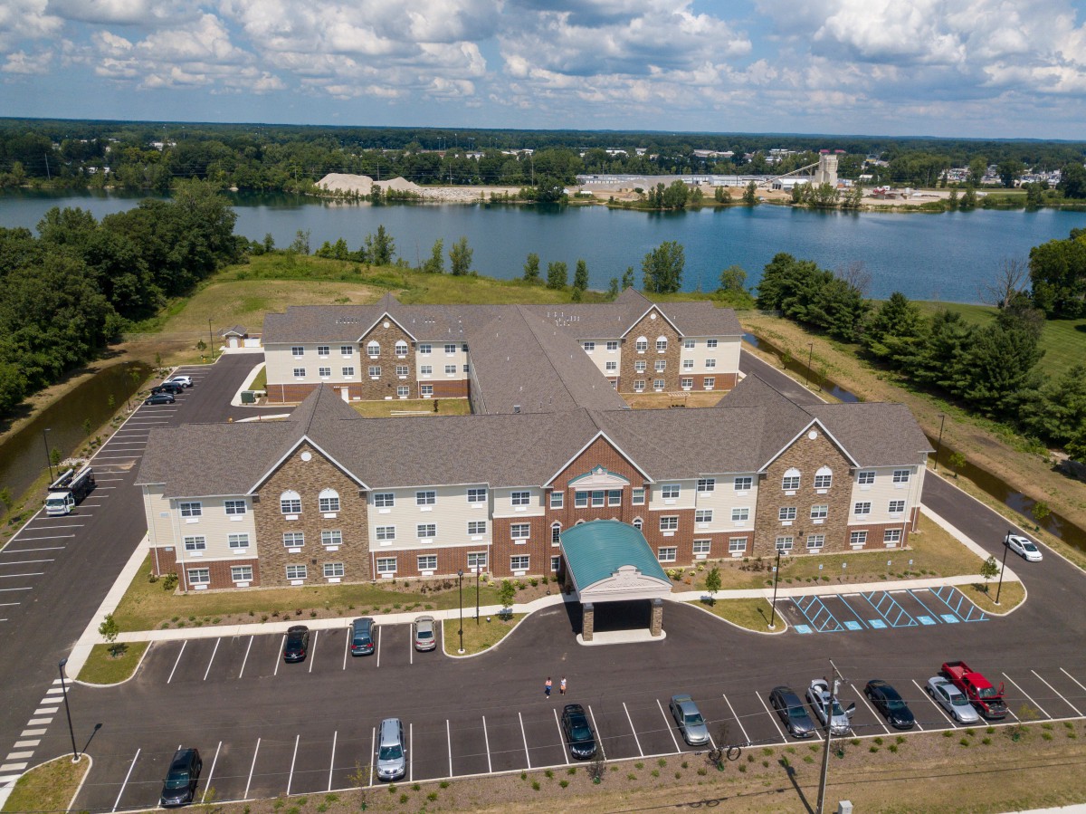 Aerial view of a large building with a parking lot, set against a scenic backdrop of a lake and cloudy sky.