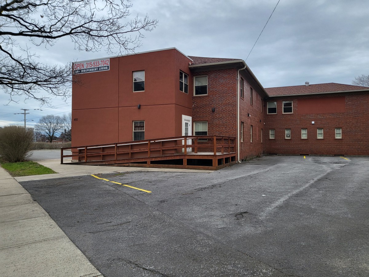 A two-story brick building with a ramp leading to the entrance, adjacent to a parking lot under an overcast sky.