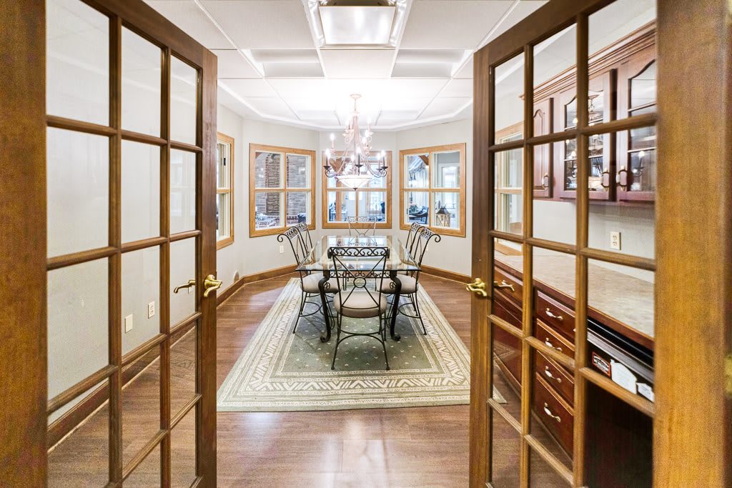 A cozy dining room with French doors, featuring a wooden table, chairs, and a chandelier, complemented by large windows and a patterned rug on hardwood floors.