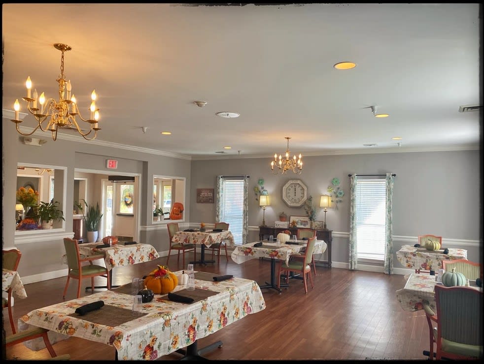 A cozy dining room with multiple tables and chairs, elegantly decorated with floral tablecloths and illuminated by chandeliers.