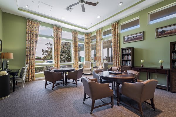 A cozy interior space with tables and chairs set up near large windows, featuring green walls and bookshelves.