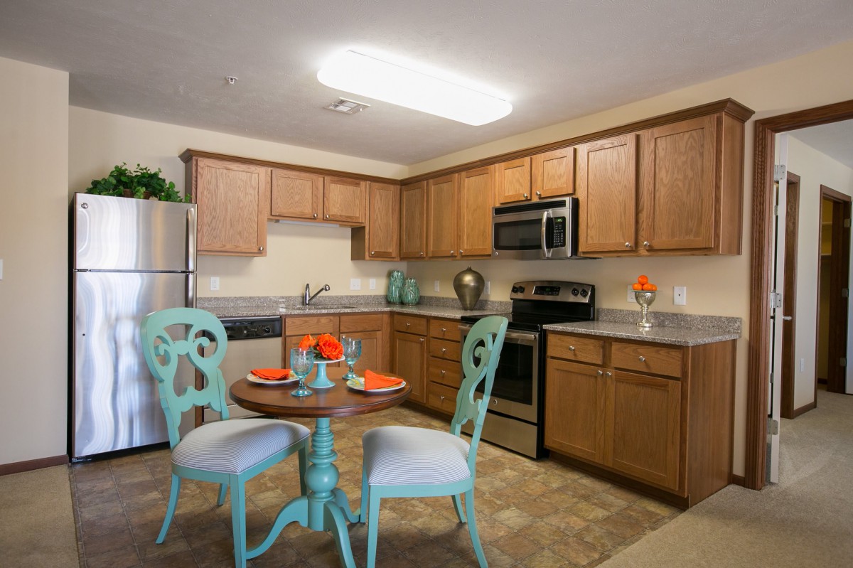 A modern kitchen with wooden cabinets and stainless steel appliances, featuring a small dining table with decorative chairs.