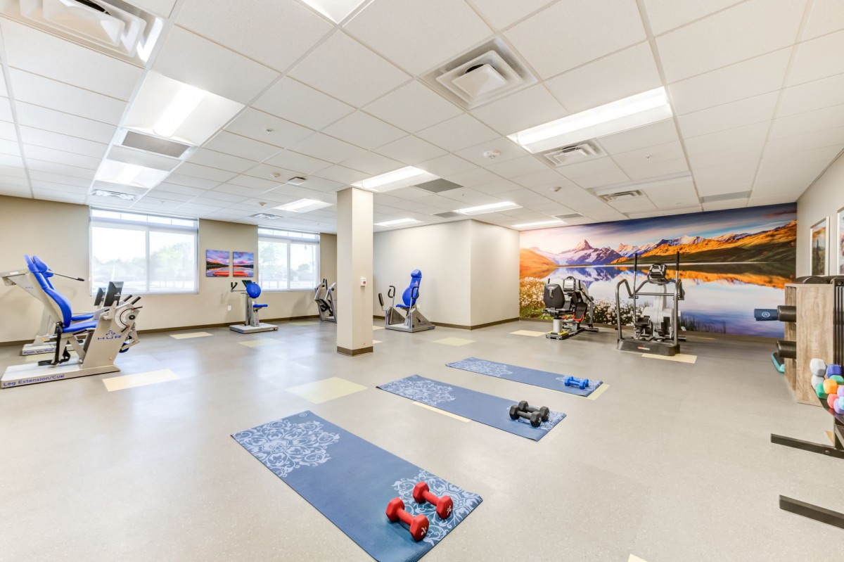 A bright and spacious gym room equipped with yoga mats, dumbbells, and exercise machines.
