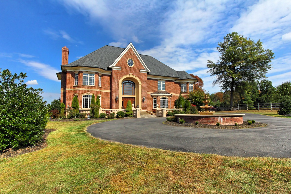 A large brick luxury mansion with a circular driveway and landscaped front yard under a partly cloudy sky.