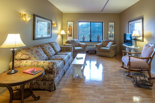 A cozy living room with floral patterned furniture and warm lighting, featuring a view through a large window.