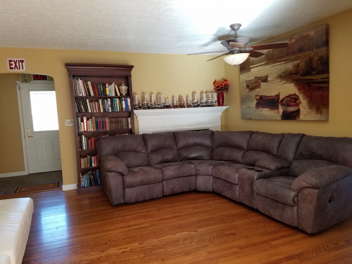 A cozy living room with a large sectional couch, bookshelf, and ceiling fan.