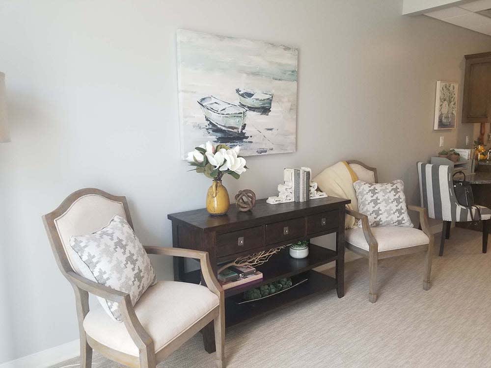 A cozy living room corner featuring elegant chairs, a dark wooden console table, and a nautical-themed painting on the wall.