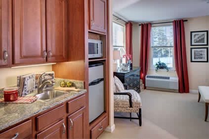 A cozy living space featuring a kitchenette with wooden cabinetry, equipped with a microwave and refrigerator, alongside an adjoining seating area with an armchair by the window.