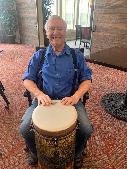 A man smiling while sitting and playing a drum indoors.