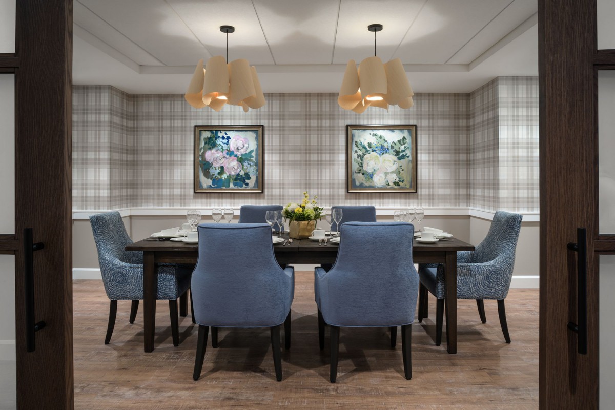 A modern dining room featuring blue upholstered chairs and a wooden table, illuminated by stylish hanging lights and adorned with floral paintings.
