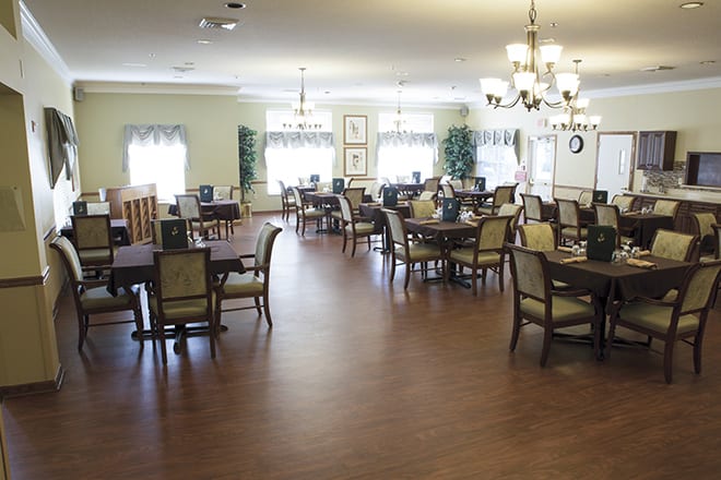 A spacious and well-lit dining room with multiple tables and chairs, featuring chandeliers and a wooden floor.