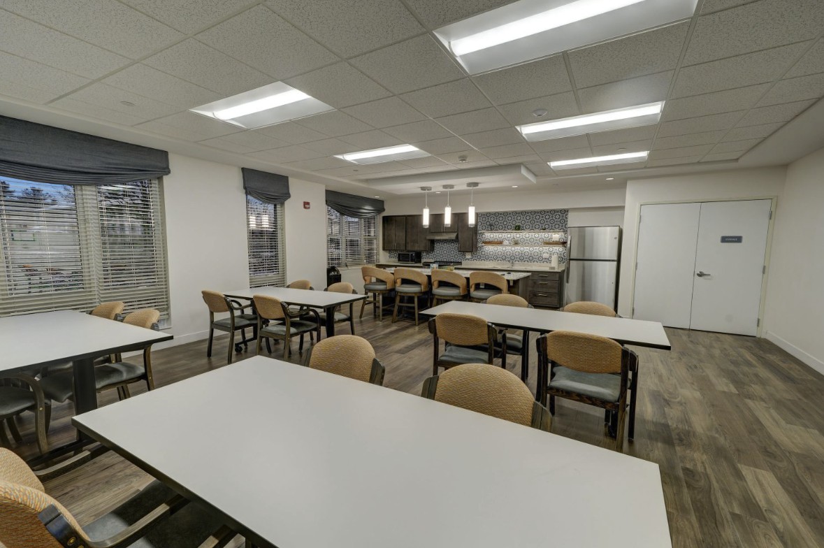 A modern cafeteria with neatly arranged tables and chairs, featuring a kitchen area in the background.