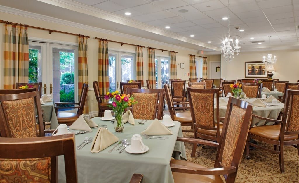 An elegant dining room with neatly arranged tables and chairs, complete with floral centerpieces and soft lighting from chandeliers.
