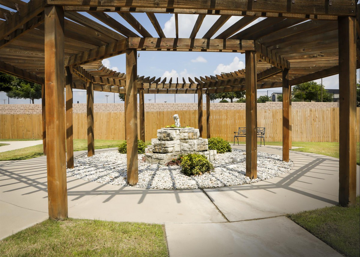A wooden pergola with a stone fountain at its center, surrounded by a landscaped garden.