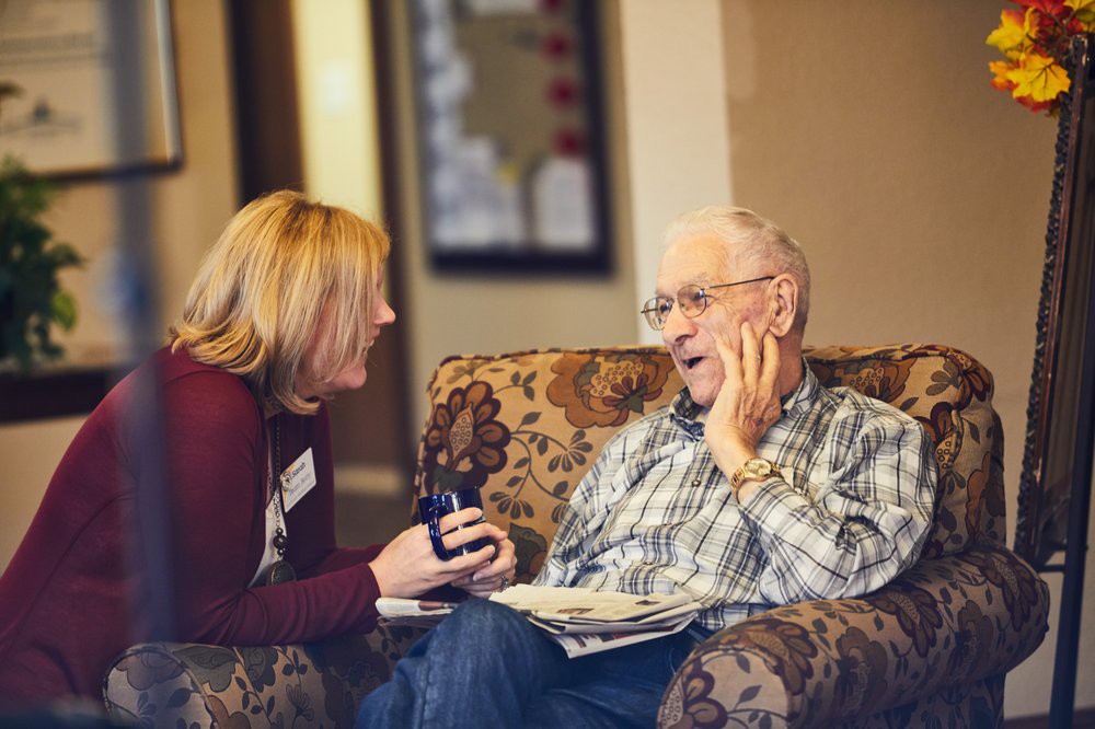 A caregiver and an elderly man share a joyful conversation indoors.