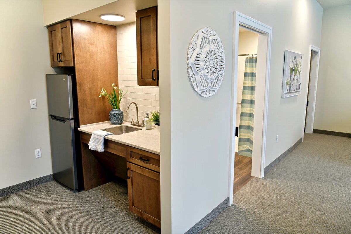 A modern kitchenette with wood cabinetry and a small fridge, leading into an adjacent room.