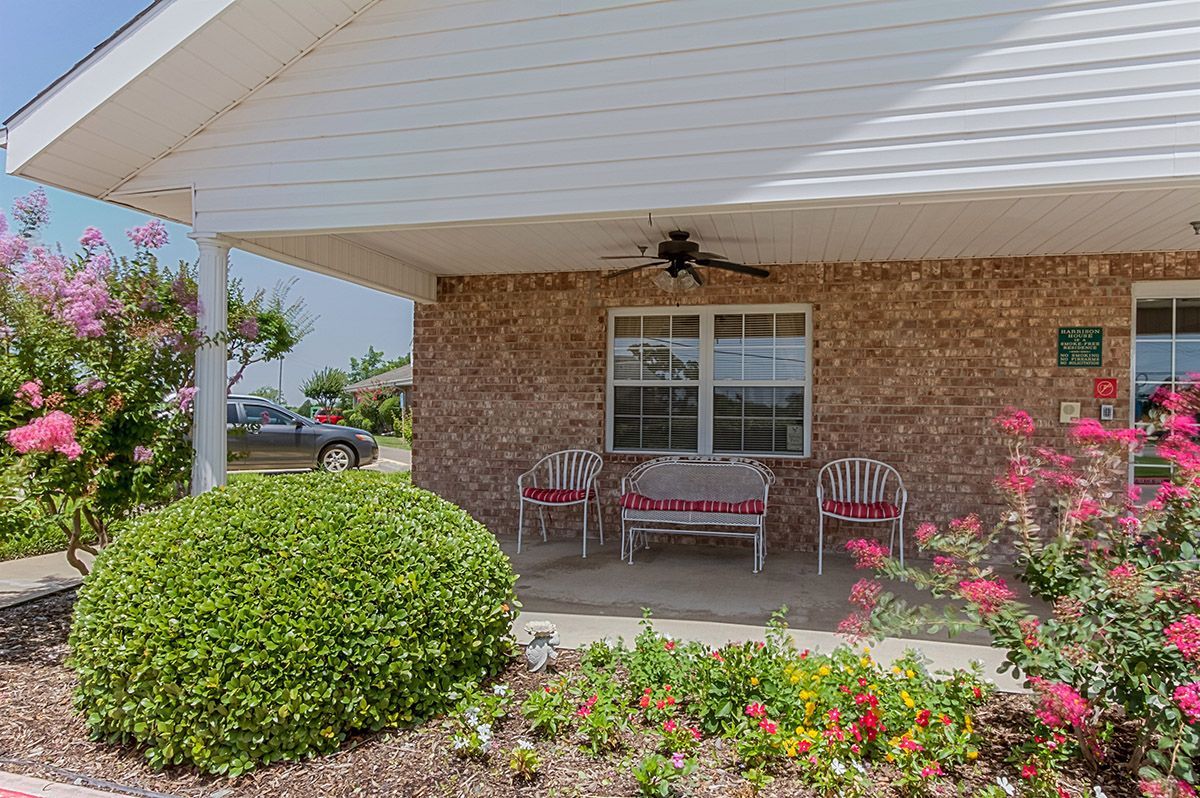 A cozy porch with two chairs and a fan, surrounded by a garden of colorful flowers and bushes.