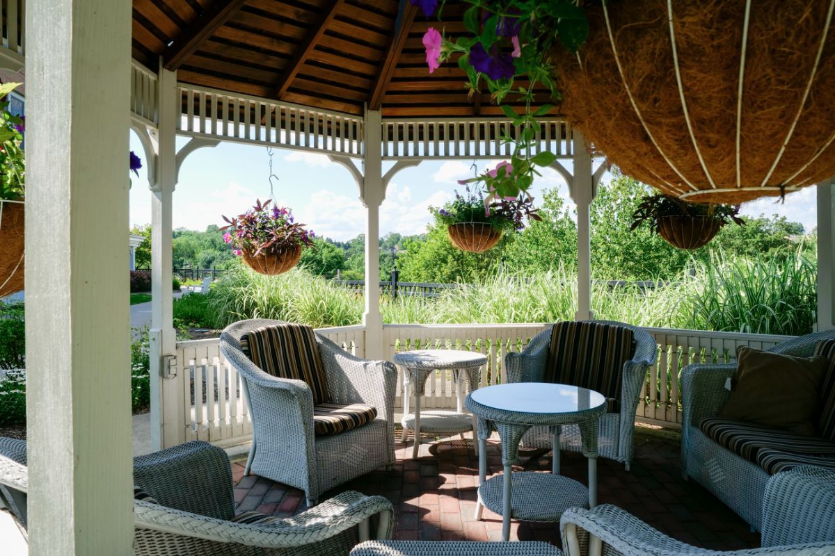 A cozy gazebo with wicker furniture and hanging plants, offering a relaxing garden view on a sunny day.