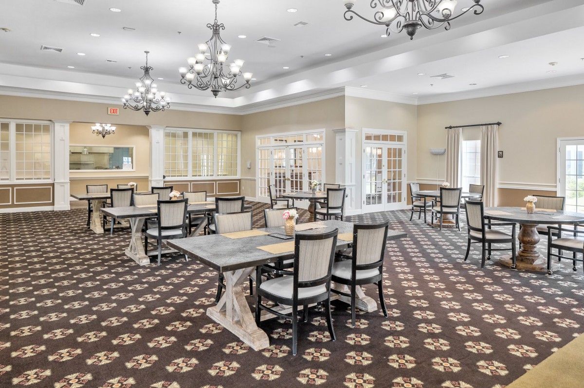 A spacious dining room with elegant chandeliers, multiple tables, and chairs arranged on a patterned carpet.