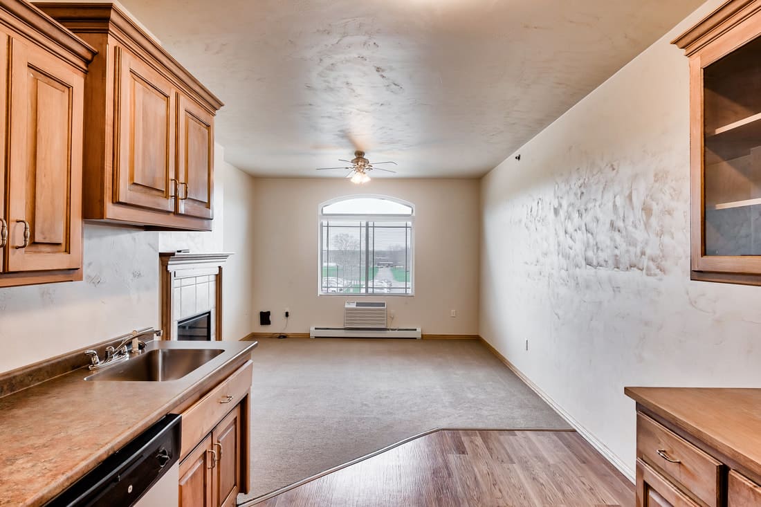 A view of an apartment's living area with a kitchen, carpeted floor, and a window allowing natural light.