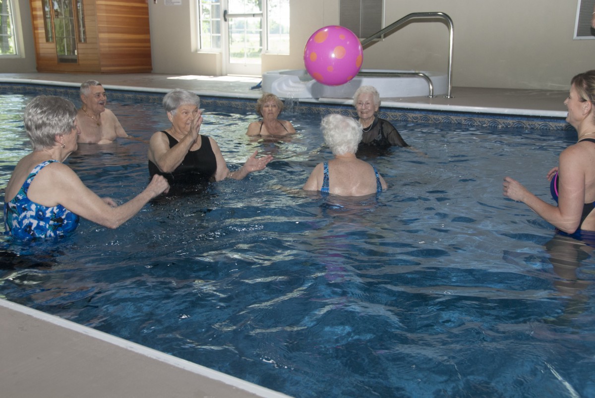 A group of seniors participating in a water exercise class, engaging with a pink ball in the pool.