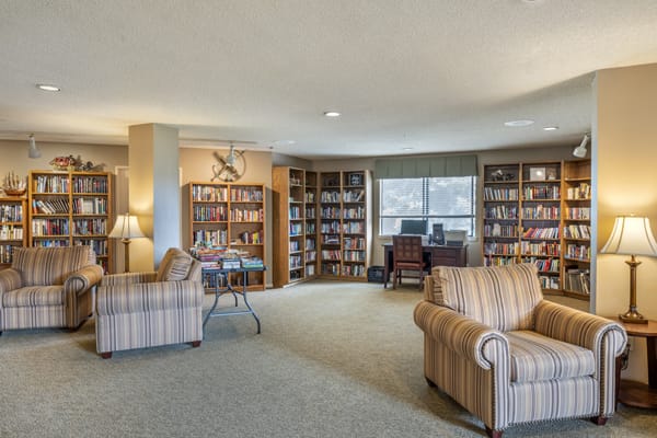 A cozy library with bookshelves, armchairs, and a reading area near a window.