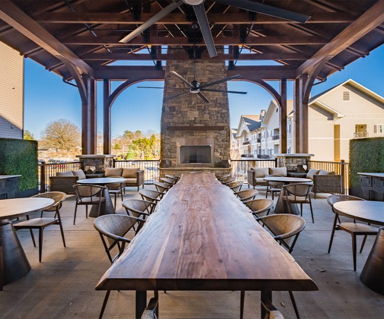 A spacious outdoor seating area featuring a long wooden table and chairs, set under a covered patio with a stone fireplace.