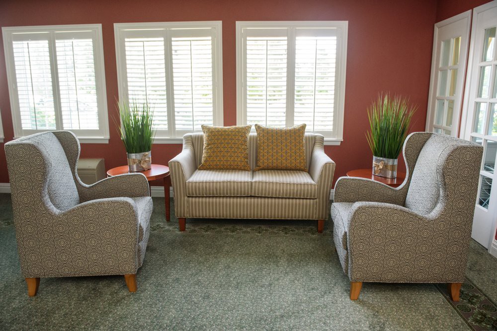 A cozy living room setup featuring a central sofa, two armchairs, side tables, and potted plants against a red wall with large windows.