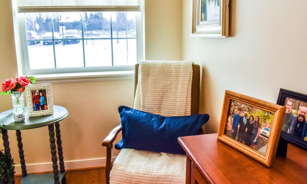 A cozy corner featuring a chair with a blue pillow by a window, illuminated by natural light, and accompanied by a framed photograph on a table.