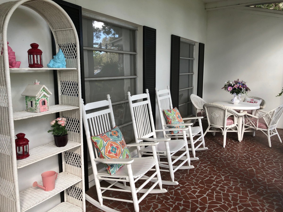 A charming porch featuring white rocking chairs with colorful pillows, decorative shelves, and a table adorned with flowers.