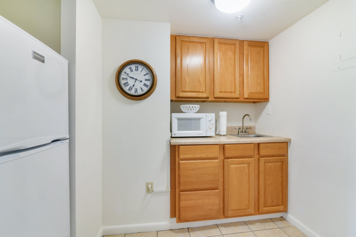 A small kitchen area with wooden cabinets, a microwave, sink, and a clock on the wall.