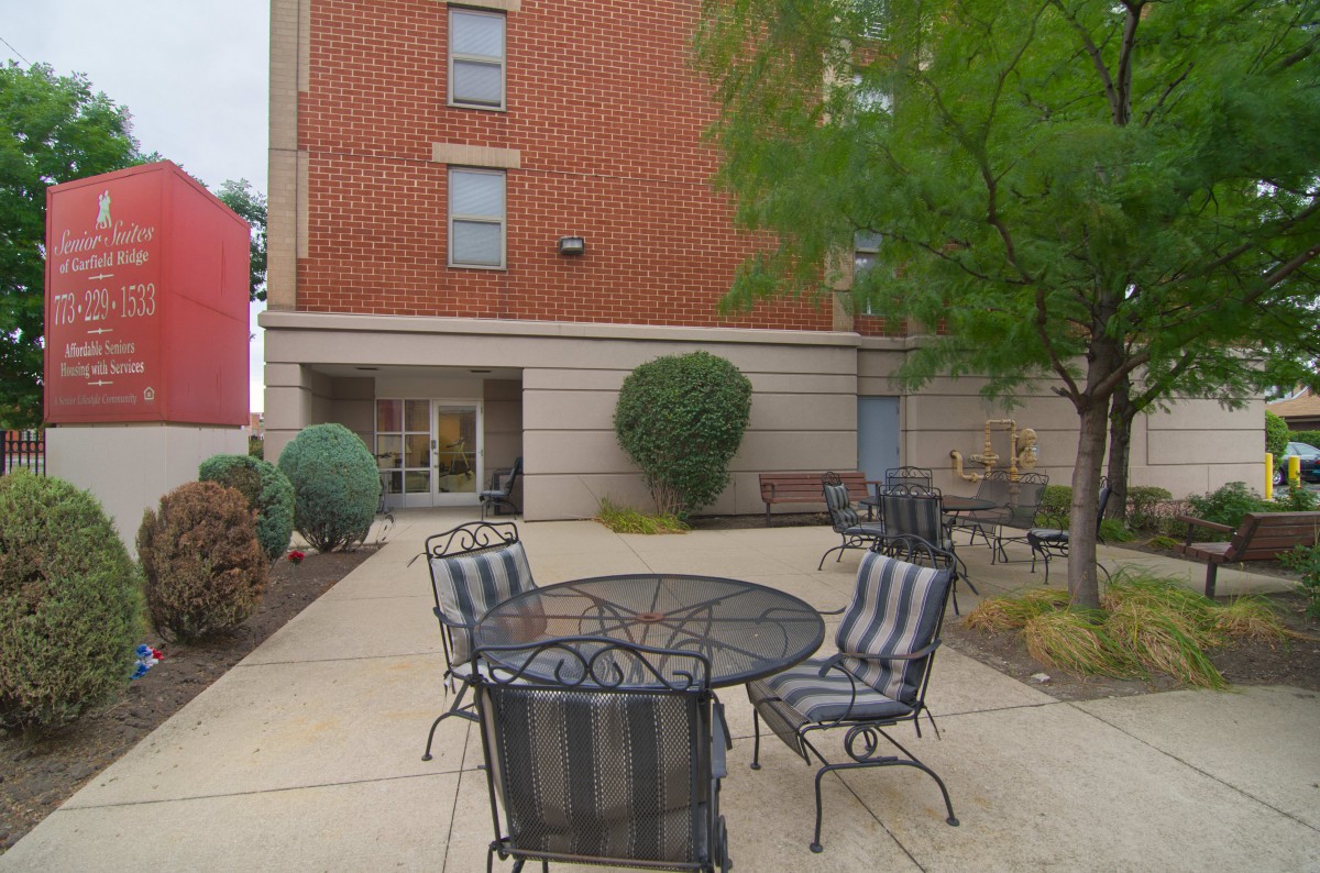 A cozy outdoor patio with metal furniture situated near a brick building, surrounded by trees and landscaping.