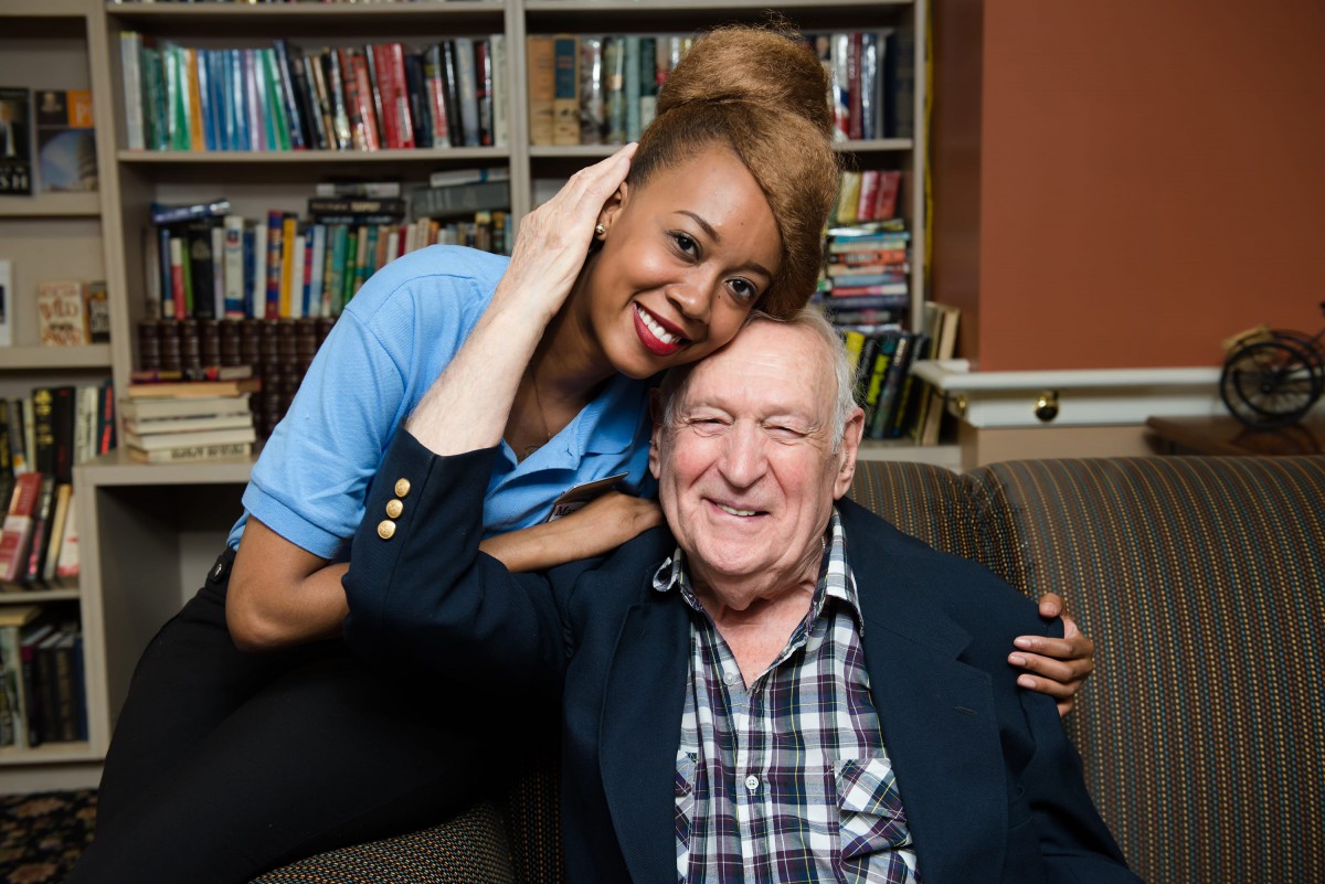 A young woman and an elderly man smiling together in front of a bookshelf.