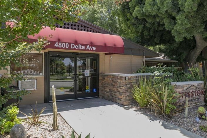 Entrance of a building with red awning and address '4800 Delta Ave', surrounded by landscaping.