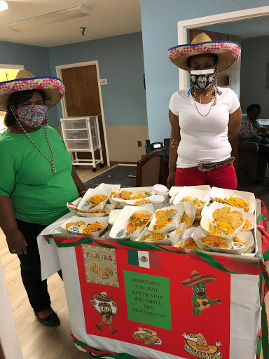 Two women wearing festive hats stand behind a table filled with delicious Mexican food at a community gathering.