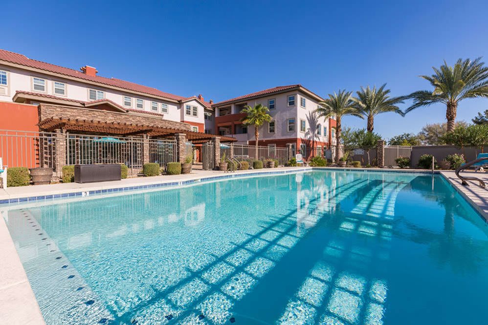 A sunny outdoor pool area with buildings and palm trees in the background.