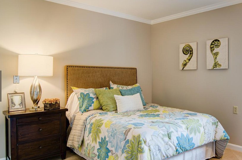 A cozy bedroom with a neatly made bed, featuring colorful bedding and pillows, a bedside lamp, and a decorative painting on the wall.