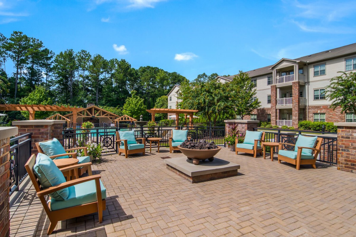 A spacious outdoor patio area with comfortable seating and a central fire pit, surrounded by lush greenery at an apartment complex.