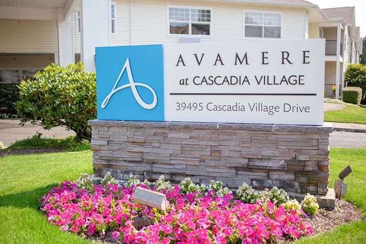 A landscaped area with colorful flowers in front of a sign for Avamere at Cascadia Village, displaying the address.