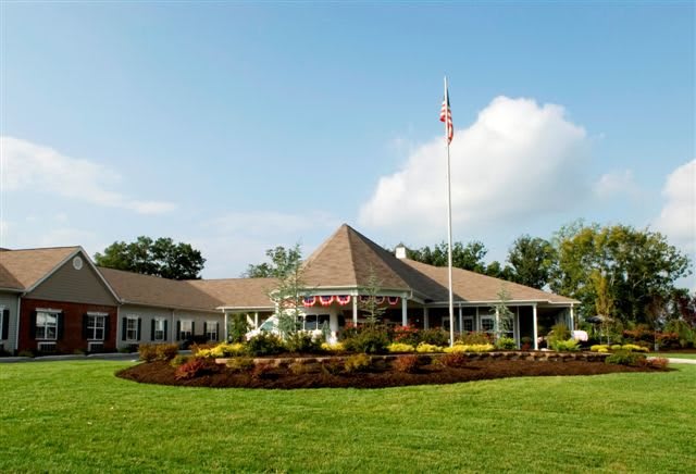 A large building with a sloped roof, surrounded by a well-maintained lawn and gardens, featuring an American flag in the foreground under a clear sky.