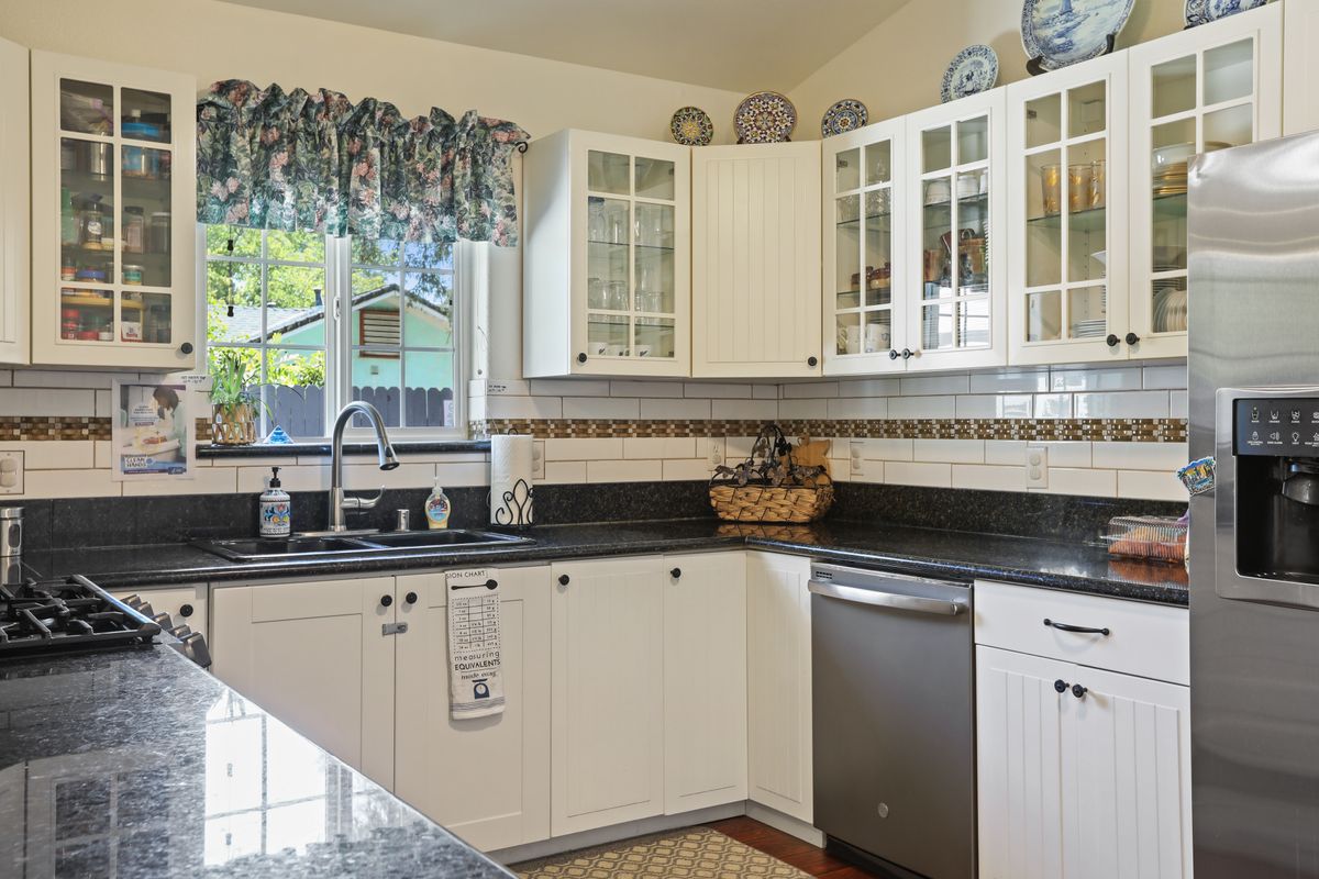 A bright kitchen with white cabinets, a black countertop, and a large window adorned with decorative curtains.
