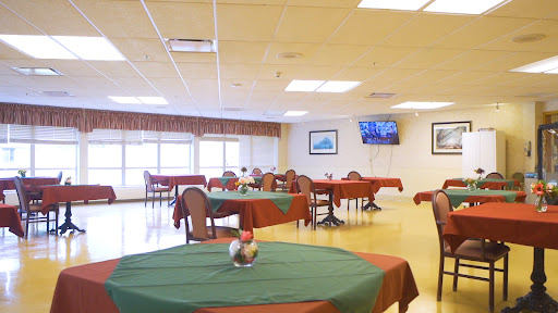 A spacious dining area set with green and red tablecloths, featuring multiple tables and chairs, illuminated by natural light.