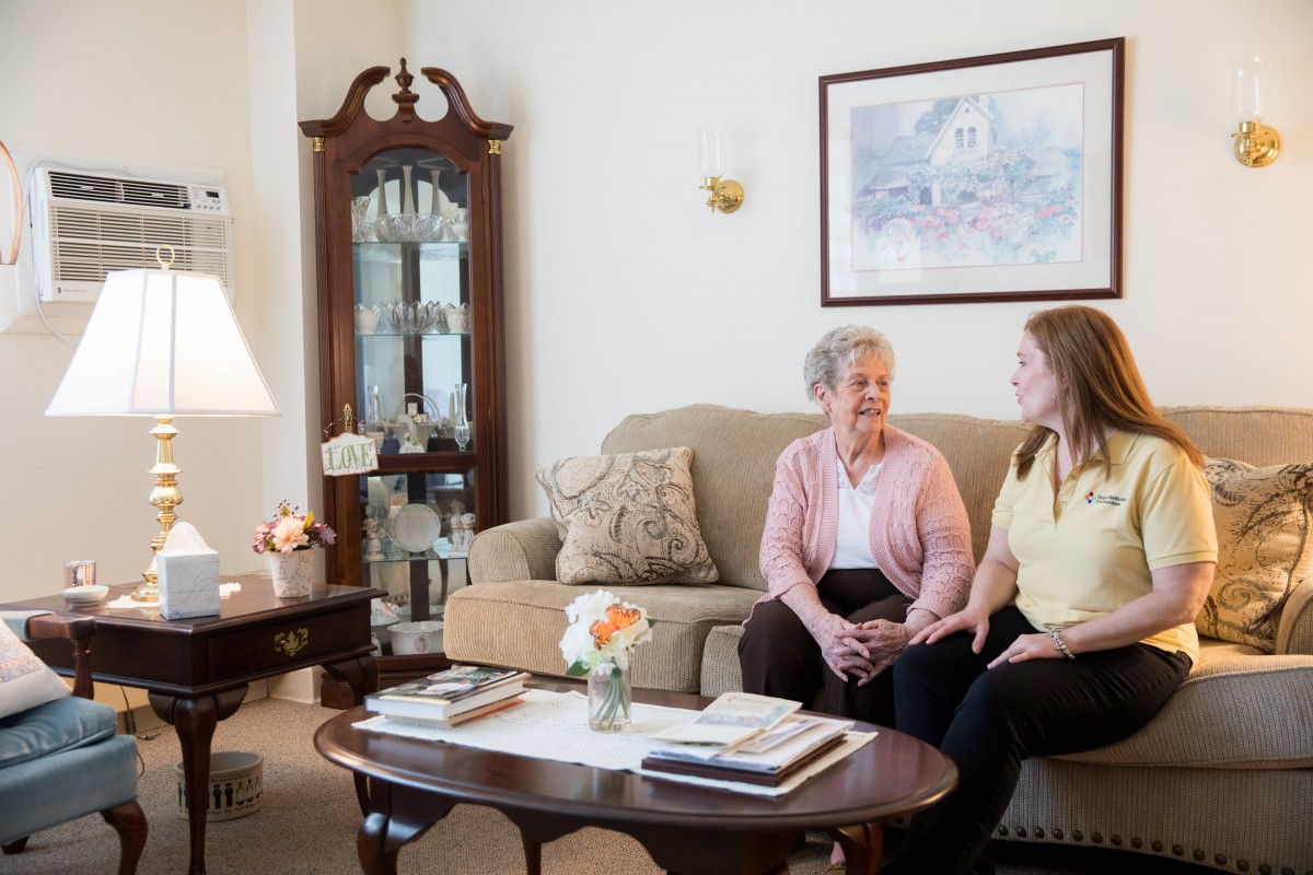 An elderly woman and a caregiver sitting on a couch in a cozy living room, engaged in conversation.