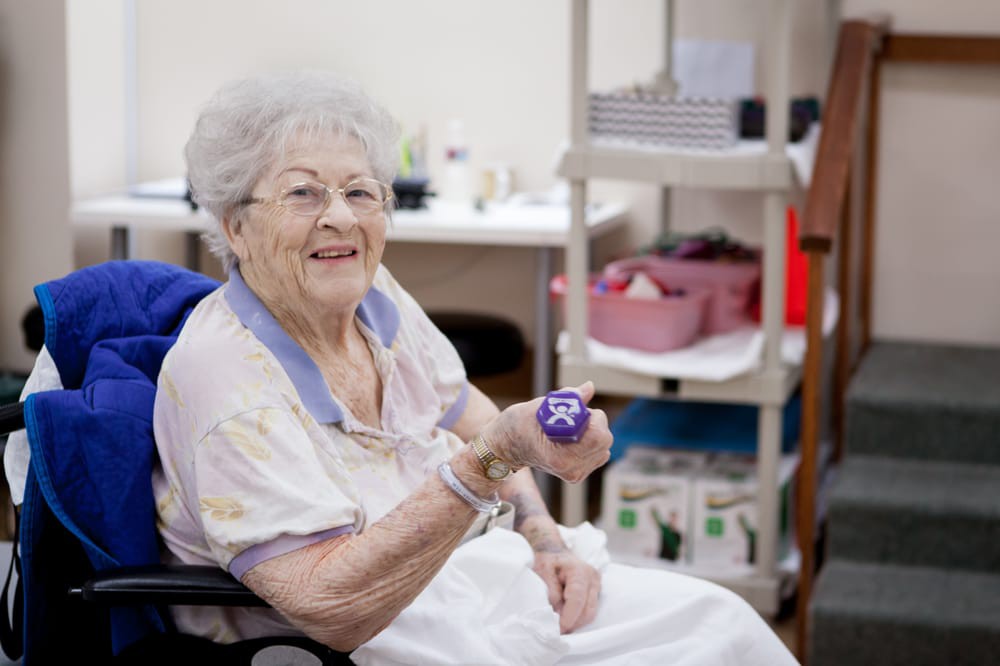 An elderly woman smiling while holding a small dumbbell indoors.