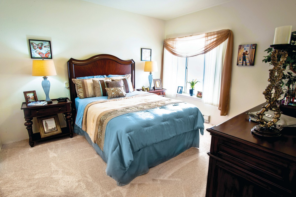 A cozy bedroom with a wooden bed frame, blue bedding, and a sunlit window.