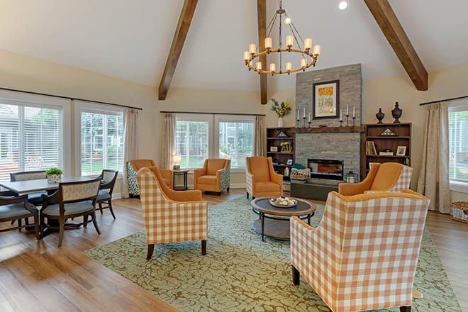 A cozy living room with a high ceiling featuring wood beams, comfortable armchairs, a fireplace, and large windows letting in natural light.