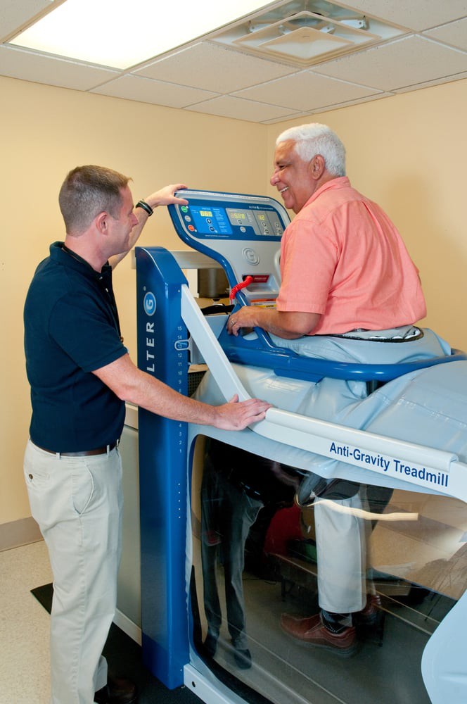 An elderly man using an anti-gravity treadmill with assistance from a trainer.